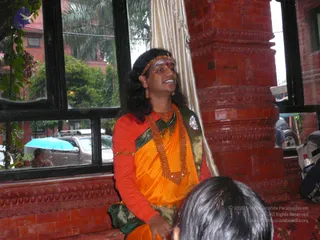 A photograph taken indoors beside a window framed by a red brick column with carved ornamentation. A person in an orange saffron-coloured garment with a green and gold border, a red fitted top, and a long beaded orange necklace stands speaking and smiling, wearing a beaded orange headband, earrings and a red bindi. Outside through the window, a light blue umbrella and parked cars are visible. The back of another person's head is in the lower foreground.