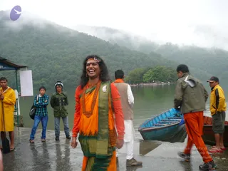 A laughing man with long dark hair stands on a wet lakeside landing in the foreground, wearing an orange blouse and an orange-and-green sari-style wrap with a green border, a red-beaded mala necklace, a red forehead mark and a decorative headband. Behind him, a blue wooden boat sits at the water's edge while men in rain jackets, one in yellow with a paddle, watch. Misty forested hills rise across the lake under overcast sky.