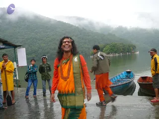 A laughing man in an orange and green ceremonial dhoti-style garment with an orange long-sleeved top stands on a wet lakeside stone landing in foggy hills. He wears a red-orange headband, a red bindi, a long red beaded mala and an ornate silver pendant. Behind him a man in an orange lower garment and dark jacket steps near a blue wooden rowboat, and to the right a man in a yellow jacket stands by a small wooden boat. On the left a man in a yellow raincoat holds an oar, a young man in a blue checked shirt and a helmeted man in a green jacket watch. A small booth with a blue sign and a blank white board sits at left, with forested hills and a wooded island on the lake behind.