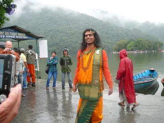 Outdoor photo beside a misty lake with forested hills behind. In the foreground a smiling person with long dark hair, a red bindi and a floral headpiece stands in a saffron-orange top and a saffron and green patterned sari-style wrap, with a heavy orange beaded necklace and a white flower at the shoulder. Behind them a man in orange robes and several people in jackets and jeans stand on a wet concrete landing. A figure in a red raincoat walks past a blue wooden boat. A ticket counter booth with a blue sign stands at left, and a small island with trees sits across the water. The photo is taken from a camera held by a hand at the lower left.