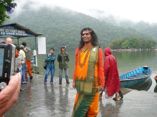 A photograph of a woman standing on a wet stone lakeside landing, smiling, with long dark wavy hair and a red-orange bindi-like mark and headband. She wears an orange blouse, a bright orange saree with a green and gold border, a heavy rust-coloured beaded necklace and a white flower brooch. Behind her a blue wooden rowing boat rests by the water, a person in a red raincoat walks past, and a ticket counter kiosk with a blue sign stands at left. Misty green forested hills rise across the lake, and several onlookers in jackets and jeans watch from the left, one photographing with a digital camera held near the frame edge.