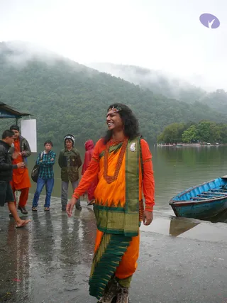 Outdoor photo beside a misty lake with forested hills behind. A man in the centre smiles broadly wearing an orange kurta-style top, an orange and green patterned wrap, a dark green-bordered shawl and a long red-brown beaded necklace, with long dark hair and a decorative headpiece. He wears sturdy laced boots and stands on a wet concrete landing next to a blue wooden rowboat. Several onlookers in jackets, a helmet and a red raincoat stand nearby, and a small roofed shelter with a white sign is at left. Appearance-based identification is not possible here; the person is described by role and attire only.