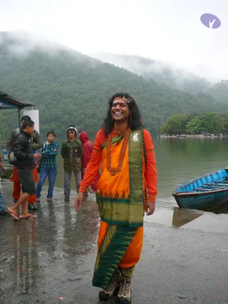 Outdoor photo on a wet stone landing beside a misty forested lake with a blue wooden rowboat at right. A smiling man with long dark hair, a red bindi and a decorated headband stands centre-frame wearing an orange long-sleeved top, a bright orange draped garment with a green and gold bordered shawl, and a thick red-orange bead necklace with a silver pendant. He wears brown hiking boots. Behind him, several onlookers in jackets, a red raincoat and a helmet stand near a small roofed kiosk. The scene is damp and overcast.