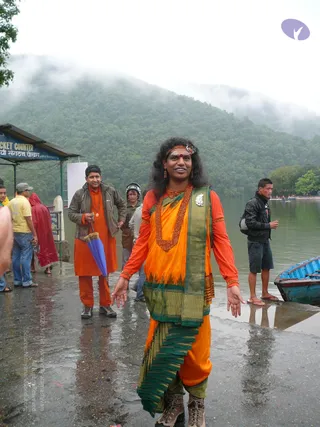 Outdoor photo on a wet concrete lakeside landing with misty forested hills behind. In the foreground a smiling person with long dark hair under an ornate headband and red bindi wears an orange and green sari-style garment, a large rudraksha-style mala necklace, beaded earrings and a white flower brooch, with arms outstretched and lace-up boots. Behind on the left a man in an orange kurta with a rudraksha mala and a grey jacket holds a blue umbrella. Other people stand nearby, including a man in a black jacket by a blue boat and a rider in a helmet. A small ticket counter sign reads Ticket Counter on the left.