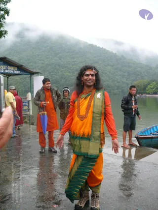 A rain-wet lakeside landing with a ticket counter kiosk and misty green hills behind. A central figure in orange and green silk draped garments, long dark swept hair, a red bindi, a headband, a wide rudraksha-style bead necklace and orange earrings walks forward with arms slightly out. Behind him a man in an orange kurta holds a blue umbrella, a man in a dark jacket and helmet stands near, and a young man in a black jacket stands by a blue boat. Outstretched hands and bright saffron clothing dominate the frame.
