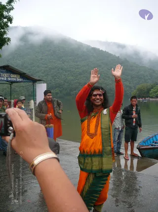A photograph at a lakeside ticket-counter landing under misty green hills. In the centre a smiling figure with long dark swept-back hair, a red bindi, a floral headpiece, orange and green ceremonial dress, a thick rudraksha mala and a large ceremonial necklace raises both hands in greeting. A photographer's outstretched arm holding a compact camera with a gold bracelet occupies the lower left. Behind, a man in a grey jacket over an orange robe holds a blue umbrella, and two young men stand near a blue wooden boat at the water's edge. A blue sign reads Ticket Counter. a person cues: long swept-back hair, ornate orange and green garments, rudraksha mala and ceremonial necklace, with the figure being greeted by a crowd.