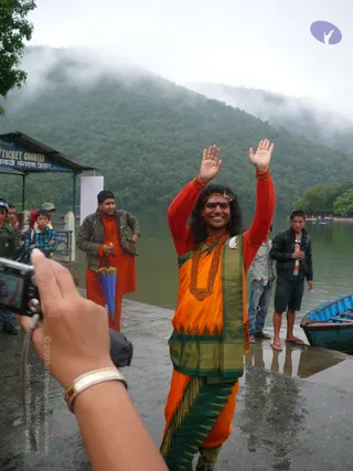 A photograph taken outdoors beside a misty lake with forested hills behind. In the centre, a smiling person with long dark hair and a red tilak on the forehead raises both hands in greeting. They wear a bright orange and red top with gold-toned armbands, an orange and green garment with a green border, a large ornate red-and-gold beaded necklace and a decorative brooch. Behind on the left, a man in an orange kurta holds a blue umbrella beside a ticket counter, and other men stand by the water's edge where a blue boat is moored. A hand holding a camera is visible at the lower left.