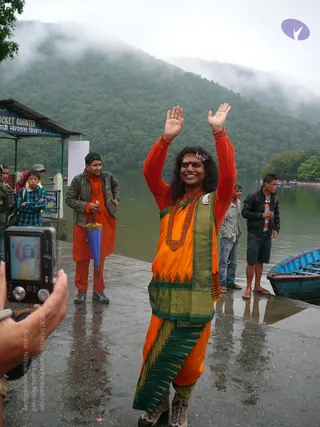 Outdoor photo on a wet lakeside stone landing beside a misty green hillside. A central figure with long dark hair, a red tilak mark and a flower headpiece stands with both arms raised and palms open, smiling. He wears a red long-sleeved top, an orange and green-bordered sari-style garment draped over the shoulders, and a thick red-beaded mala with a white floral ornament, plus hiking boots. Behind him a man in an orange kurta and brown jacket holds a blue umbrella and smiles, a boy in a plaid shirt stands near a ticket counter sign, and a man in a black jacket holds a bottle beside a blue wooden boat. In the foreground a hand holds a digital camera showing the figure on its screen.