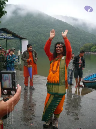 A smiling figure with long dark hair, a red bindi and a flower headpiece stands on a wet stone lakeside landing with both palms raised overhead. He wears a red long-sleeved top, an orange and green sari-style wrap, a thick red rudraksha-style bead mala, a white flower pendant and hiking boots. Behind him, men in orange kurta-style clothing and jackets watch, one holding a blue umbrella, beside a blue wooden boat and a ticket counter sign on a misty green hillside. A person in the lower left photographs him with a digital camera showing a similar image on its screen.