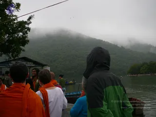 A misty, forested hillside rises above a lake at a boat-ticket counter on the shore. In the foreground, several people stand on a wet landing: a figure in a black and green hooded rain jacket faces the water, a person in a white top with an orange shawl stands beside a bald man in an orange robe, and a figure in an orange wrap on the left faces away. A standing person in green with an orange garland and a headpiece looks toward the group. Small wooden boats with oars are moored along the water.