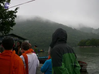 A misty lakeside scene with forested hills rising into low cloud. In the foreground, several people stand at a wooden boat landing: a man in a black and green rain jacket with hood up, a figure in a blue jacket, a person in a white top with an orange shawl, and others in orange shawls. A small ticket counter shed stands at left, and wooden rowboats float on grey-green water, with a rowing boatman in green in the middle distance.