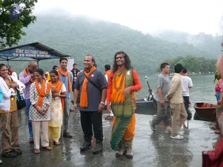 Outdoor photo on a wet lakeside landing beside a misty forested hillside. Beside him stands a man in a grey shirt, black trousers and boots, with an orange scarf. Other people in orange shawls stand on the left by a blue sign reading Ticket Counter, and wooden boats rest on the water behind.