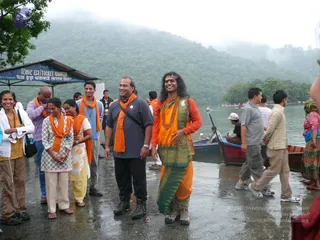 Outdoor photo at a misty lakeside with wooded hills behind and a wet stone landing. A central figure in an orange top, green-and-gold draped garment and orange dhoti-style lower wrap, wearing a long dark swept-back hairstyle, red tilak mark and a large beaded orange-brown mala, stands smiling beside a man in a grey shirt with an orange scarf. Around them, several people wear orange shawls, including a woman in a white and grey outfit and a woman in a yellow salwar. Wooden rowboats are tied at the shore and a ticket counter sign stands at left.