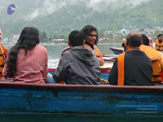 A photograph of a group seated in a blue wooden boat on a misty lake with forested hills and a town behind them. Several people wear orange and yellow life vests. In the centre, a man with long dark hair and a red tilak on his forehead smiles, wearing an orange-trimmed vest. In the foreground, a woman with long dark hair wears a red striped shirt, a man in a grey hooded jacket sits with his back turned, and a bald man in a black top with an orange vest sits at right.