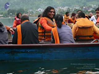 A photo of a group riding in a wooden boat on a lake with a hilly town in the background. Most passengers wear orange life vests over their clothes. In the centre, a smiling man with long dark hair and a red-orange garment, with a red headband and a red mark on his forehead, sits turned toward the people beside him; he is a person by appearance, with long swept-back hair and a distinctive orange and red clothing combination, though the cues are limited. Other passengers include a woman in a grey top with her hair tied back, a man in a dark jacket seen from behind, and a woman in a red life vest at left. The boat has a dark blue hull with a red rim, and the water is grey-green.