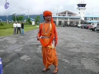 A photograph of a young man standing on a paved airport apron in front of a low terminal building with a control tower. He wears a saffron-orange turban, an orange sleeveless-style kurta over orange dhoti-style drapery, and a long orange shawl held in one hand. He has layered rudraksha-style beaded malas and a gold ceremonial necklace, a patterned waist sash, and orange sandals. Behind him, three men in white or dark shirts stand on grass beside a fence, and parked cars and motorcycles line the lot. Overcast hills rise in the background. The figure has distinctive saffron attire and beaded malas, but his appearance is not conclusive enough to confirm a specific identity, so he is described by role only.