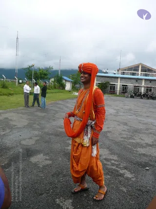 A photograph of a smiling man standing on a paved open ground in front of a multi-storey building with a blue-trimmed roof and parked motorcycles. He wears an orange turban with the tail draped over his left shoulder, an orange top with gold-trimmed sleeves, a long orange shawl, a beaded necklace, a striped white and orange sash tied at the waist, orange dhoti-style trousers and orange-and-black sandals. Behind him, two men in white shirts and trousers stand on grass beside a third person in dark clothing, with radio masts and misty green hills in the background.
