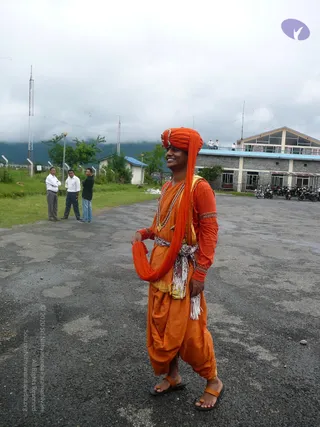 A smiling man stands on a paved open ground in front of a multi-storey building with a blue-trimmed roof, mountains and low cloud behind. He wears an orange turban, an orange long-sleeved kurta with decorative trim, orange dhoti-style trousers, a white-and-orange sash at the waist, a layered rudraksha mala and a gold pendant, and orange sandals. Part of the orange turban cloth is draped over his shoulder and held in one hand. Three men in white shirts and trousers stand on grass to the left, and a telecom tower rises in the background. a person is not visibly identifiable from appearance here, so no candidate is named.