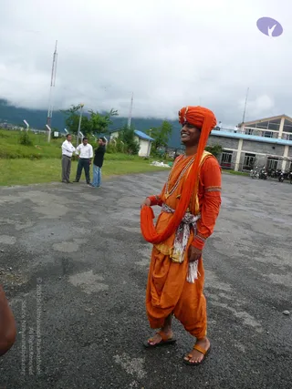 A smiling young man stands on an asphalt apron outdoors, wearing an orange turban with a long orange cloth draped over one shoulder, an orange kurta with gold-trimmed cuffs, orange dhoti-style trousers, a white and red striped sash at the waist, a beaded necklace, and orange leather sandals. Behind him, three men in shirts and trousers stand on grass beside a low building with a flat blue roof, a lamp post, communication towers, parked motorcycles, and misty green hills under a cloudy sky.