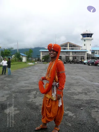 A smiling man stands on a paved airfield apron in front of a low stone terminal building with a white control tower and blue trim. He wears an orange turban with a small ornament, an orange long-sleeved top with decorative trim, an orange draped wrap over one shoulder, orange dhoti-style trousers, a beaded necklace and an orange-and-white patterned sash at the waist. Behind him, parked cars and motorcycles line the lot, two men in white and dark clothing stand on grass to the left, and misty hills rise under an overcast sky.