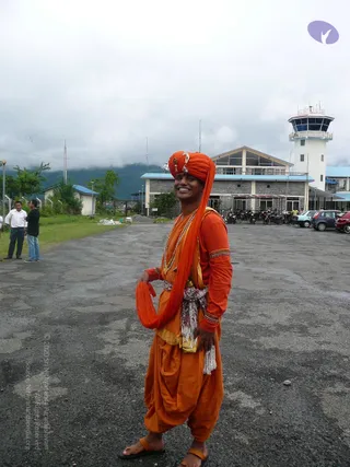 A smiling young man stands on an asphalt apron in front of a grey stone airport terminal with a white control tower and blue roof trim. He wears an orange turban with a gold ornament at the front, an orange long-sleeved tunic with gold-embroidered cuffs, a white and red sash at the waist, orange dhoti-style trousers and orange sandals. A rudraksha-style bead mala hangs at his neck, and an orange shawl drapes over one shoulder. Behind him, parked motorcycles and cars line the lot, and two men stand on grass to the left under low clouds and forested hills.