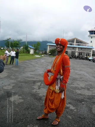 A smiling man stands on a wet asphalt apron in front of a stone airport terminal with a control tower. He wears an orange turban with a small ornament, an orange long-sleeved top, an orange dhoti-style lower garment, a white and orange waist sash, and a long orange shawl draped over one shoulder, held in one hand. Orange sandals are on his feet, and a rudraksha-style bead necklace hangs at his chest. Behind him, a few men in white shirts and trousers stand on grass near a fence, a parked row of motorcycles and cars sits beside the terminal, and low cloud covers the green hills. A purple logo appears in the upper right corner.