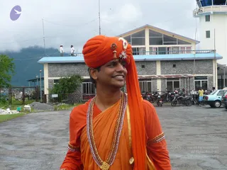 A smiling man stands on a paved open area in front of a two-storey stone-and-concrete building with a blue-edged balcony and a white control tower at right. He wears a bright orange turban with a decorative gold-and-red brooch and an upright white ornament, a long orange sash falling over his shoulder, an orange robe with gold-trimmed sleeves, and a layered multi-strand beaded necklace with a gold pendant. Parked motorcycles, a light blue car and two people on the balcony appear behind him under a cloudy sky with forested hills. Printed copyright text and a logo overlay the lower right and upper left corners.