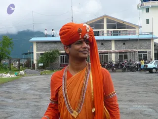 Outdoor photo on a paved forecourt in front of a two-storey stone building with a blue-trimmed balcony and parked motorcycles and a small car to the right. A man in the foreground wears an orange-red turban with gold and jewelled ornaments and a small red mark on his forehead. He is dressed in orange-saffron robes with a gold-bordered sash, layered beaded and gold necklaces, and a long orange scarf draped over one shoulder. He smiles and looks off to the side. Misty green hills rise behind the buildings, and a faint purple logo sits in the upper left corner.