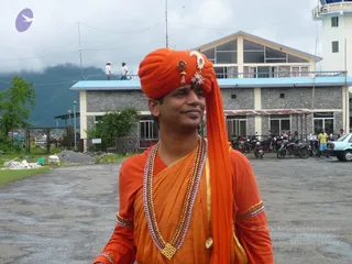 Outdoor photo on a paved courtyard in front of a two-storey stone building with a blue-trimmed roof, parked motorcycles and a white car at right, and misty green hills behind. A man in the centre wears a bright saffron-orange turban with a jewelled gold ornament at the front, a saffron-orange robe with gold-trimmed sleeves, and layered beaded and crystal necklaces. He smiles and looks off to the side. Two people stand on the rooftop balcony behind and another figure stands at right.
