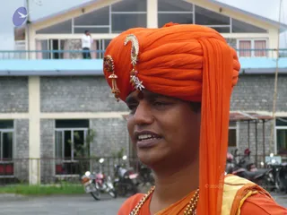 Close photograph of a smiling man in an orange turban wrapped in a long orange cloth draped over his shoulder. The turban is decorated with a gold and jewelled ornament with red stones and dangling tassels at the front. He wears orange robes with gold-coloured beaded necklaces and a red tilak on his forehead. Behind him stands a grey stone building with a blue-railed balcony, where a person in white stands, and several parked motorcycles are visible along a low fence on a paved courtyard.