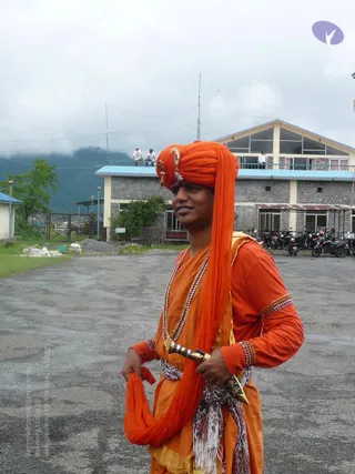 A man stands on a wet paved courtyard in front of a two-storey stone building with a blue-trimmed roof and a parked row of motorcycles. He wears an orange turban decorated with a gold ornament at the front, an orange robe and kurta with beaded bangles on his arms, and layered beaded and gold necklaces. A patterned white and brown shawl is tied at his waist, and he holds an orange cloth in one hand and a decorative metal item in the other. Two people stand on the upper balcony behind him, and cloudy hills rise in the background.