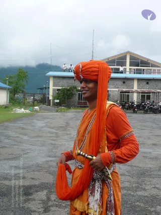 A smiling man stands on a paved lot in front of a multi-storey stone building with a blue-trimmed roof and balcony. He wears an orange turban with a gold ornament at the front and a long orange cloth draped down his back and over his arm. His orange top has beaded bangles on the upper arm and wrists, and he wears layered beaded necklaces, a spotted scarf at the waist, and holds a small brass-and-metal horn-like object. Motorcycles are parked at the right, two people stand on a rooftop, and misty green hills rise behind the building.