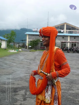 A man stands on a paved open lot in a misty hill setting, wearing a bright orange turban with a long orange cloth hanging down one side and gold-coloured ornaments at the front. He wears an orange long-sleeved kurta with beaded bangles on his wrists and armband, layered gold and beaded necklaces, an orange shawl or dhoti wrap, and a patterned white-and-grey sash at the waist with a dagger-like curved weapon tucked into it. Behind him are a grey stone building with a blue-roofed balcony, two figures on a rooftop, parked motorbikes, a small blue-roofed hut, green trees and forested mountains under low cloud.