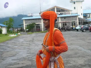 A man stands outdoors on a wet paved open area in front of a multi-storey grey stone building with blue-roofed sections, a tower and parked motorcycles and cars behind him. He wears an orange turban with a saffron-coloured long wrap draped over his shoulder, an orange kurta with decorated gold-banded cuffs, multiple beaded necklaces, and a white patterned sash at the waist, and he holds a sword-like hilt in his hand. Two people stand on a rooftop in the background, and a green tree and misty hills appear behind the buildings. The scene shows a plain outdoor location with no visible ritual activity.
