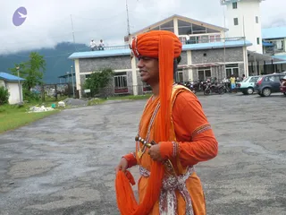 A man walks across a paved lot in a stone-and-concrete compound under overcast skies. He wears an orange turban with a long orange wrap draped over his shoulder, an orange kurta with embroidered beaded cuffs, and a white patterned sash at his waist. Several strands of beads hang around his neck and a dagger-style item is tucked at his waist. Behind him are a multi-storey building with blue-trimmed roofs, parked motorcycles and cars, and a grassy strip with a small tree.
