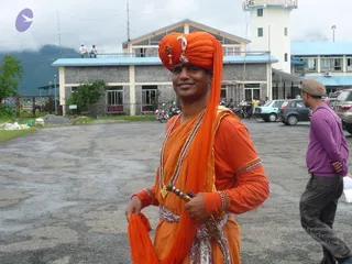 A smiling man in a saffron-orange turban with a jewelled crescent ornament stands on a paved lot in front of a multi-storey building with a light-blue roofline and a control tower. He wears orange robes with gold-embroidered cuffs, a gold bead necklace and a rudraksha-style mala, and holds a dagger-like sheathed blade at his waist with an orange cloth draped over his arm. Behind him, another man in a purple long-sleeved top and cap walks past parked cars and motorcycles on a cloudy day, with two people seated on a rooftop and green hills in the background.