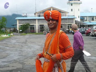 A smiling man stands on a wet paved apron in front of a grey stone airport terminal with a white control tower. He wears an orange turban decorated with a silver ornament and a red-and-gold brooch, an orange kurta with gold-banded sleeves, a long orange shawl, and layered beaded necklaces, with a curved ornamental dagger tucked into a white patterned sash. Behind him, another man in a purple sweater and grey trousers walks past with a white paper in hand, and parked cars and motorbikes line the lot.