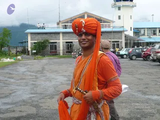 A smiling man in a bright orange turban with a gold and white ornament at the front and orange robes stands on a paved airport apron. He wears layered beaded necklaces, gold armbands, and a sword-hilted dagger tucked at his waist, and holds a corner of his orange wrap. Behind him, a grey-brick terminal building with a blue roof and a white control tower stands beside parked cars and motorcycles, with misty hills in the background. A person in a purple top walks past, and two people sit on a rooftop railing.