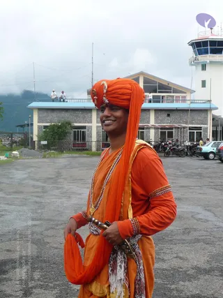 A smiling man stands on a paved airport apron in front of a two-storey grey stone building with a white control tower behind. He wears an orange turban with a crescent and jewelled ornament, an orange long-sleeved tunic and wrap with a gold-and-beaded necklace, beaded bracelets, and a leopard-print scarf tucked at the waist with a curved dagger-like hilt. Small figures stand on the roof, a few parked motorcycles and cars sit to the right, and a wooded hill rises behind under overcast sky.
