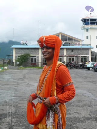 A smiling man stands on a paved airport apron in front of a stone-faced terminal building with a control tower behind him. He wears an orange turban with a gold crescent ornament and a long orange shawl draped over his shoulder, an orange kurta, a multi-strand beaded necklace and beaded armbands. A curved metal dagger is tucked into a patterned fabric sash at his waist. Parked cars and motorcycles and two people on the rooftop are visible in the background under a cloudy sky.