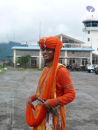 A smiling man stands on a paved airfield apron in front of a stone-faced terminal building and a white control tower. He wears an orange turban decorated with a gold ornament and a crescent-style jewel, an orange long-sleeved top, and an orange-and-gold wrap draped over his shoulder. He has layered beaded necklaces and beaded bangles on his wrists, and a curved dagger with a dark hilt is tucked into a patterned sash at his waist. Behind him, motorcycles are parked along a railing, a small white car is visible on the right, and a few people stand on a rooftop under a cloudy sky with forested hills behind.