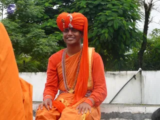 A smiling young man sits on a concrete ledge in front of a white wall and leafy green trees. He wears a bright orange turban decorated with a gold ornament and a white jewelled brooch, an orange long-sleeved top, an orange wrap with a gold-bordered edge, and a long beaded gold-toned necklace. Both hands rest on his knees, and a sliver of another person in orange clothing is visible at the left edge of the frame.