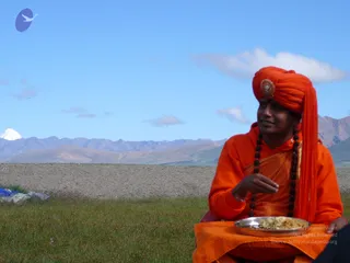 A photograph of a man seated outdoors on grass in a high mountain landscape under a clear blue sky, with a snow-capped peak visible on the left and brown ridges behind. He wears a bright orange turban with a round gold-toned brooch, an orange robe with an orange shawl, and a long dark rudraksha mala hanging to his waist. He holds food with his right hand from a silver-toned plate of rice resting on an orange cloth in his lap. A blue tarp and grey stones lie at the lower left.