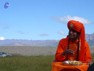 A man in a saffron-orange turban with a round brooch and a matching orange robe sits on grass in a high mountain valley under a clear blue sky. He wears a long dark rudraksha mala and holds a steel plate of food in his lap while looking off to the side. Behind him are brown mountain ridges and a snow-capped peak on the left, with gravel ground and a blue tarpaulin at the lower left. a person cues are present: a distinctive orange turban with brooch, saffron clothing and a rudraksha mala. The page does not indicate a person was at this event, so the person is recorded as an unsure candidate.