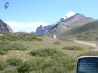 A blurred outdoor photograph of a high-altitude mountain landscape taken through a vehicle window. A snow-capped peak rises at centre-right above rugged brown rock ridges and a dry, grassy valley with low green shrubs in the foreground. A pale dirt track curves through the scene at right, and a white vehicle is parked or driving in the middle distance. The edge of a vehicle side mirror is visible at the lower right. No people are visible.