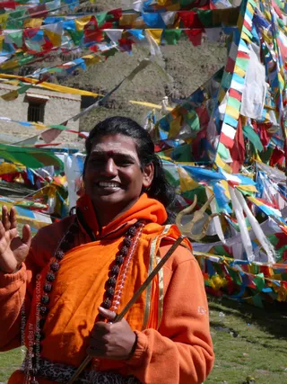 A smiling man with long dark swept-back hair and a red tilak on his forehead stands outdoors in front of a hillside building and a dense row of colourful prayer flags in red, yellow, green, blue and white. He wears an orange-saffron fleece wrap with gold trim and a long rudraksha mala of dark beads, and holds a brass trident with his left hand while raising his right palm in greeting. Green grass is in the foreground. Appearance cues are distinctive but no printed name or event is visible, so identity is uncertain and he is described by role as a sadhu-style devotee.