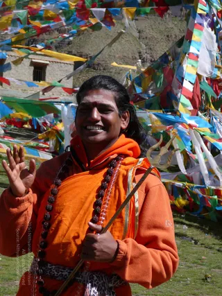 A smiling man with long dark hair, a red bindi and a dark mark on his forehead stands outdoors before a hillside building and dense rows of colourful prayer flags in red, yellow, blue, green and white. He wears an orange-saffron hooded top with gold trim and a leopard-print cloth at the waist, a long rudraksha mala hangs across his chest, and he raises one hand in greeting while holding a brass trident in the other. Green grass and a discarded can lie at his feet.