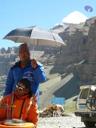 Outdoors in a rocky mountain valley, a snow-capped peak rises behind layered brown cliffs under a clear blue sky. In the foreground a seated person in an orange robe, wearing a rudraksha mala and dark sunglasses with orange lenses, raises one index finger while holding a small piece of food near a metal plate on an orange-draped table. Standing behind them, a man in a blue padded jacket with a knit cap and glasses holds a black-and-silver umbrella over the seated figure. To the right a white Toyota-style vehicle is parked beside a blue roadside sign and stacked bundles of firewood on gravel.