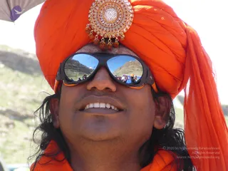 Close-up photo of a smiling man wearing a vivid orange turban decorated with a large gold and crystal brooch hung with rudraksha beads, and dark sport sunglasses with orange temple arms. His long dark hair falls over an orange shawl. Mirrored lenses reflect a mountain slope and a crowd of people. Behind him are blurred green hills and bright sky.