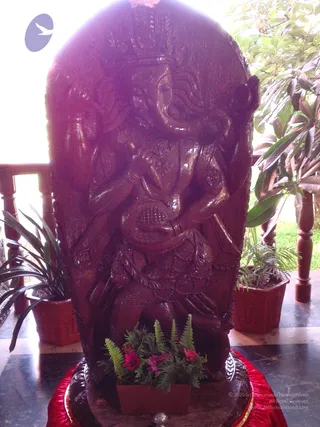 A photograph of a large dark wooden carved panel with a deity murti in relief, showing multiple arms and a crown-like headdress. The carving is set on a red cloth-draped base in front of a stand of potted plants, with small pink flowers and green fern fronds arranged at its foot. Wooden balusters and a marble floor are visible behind, with greenery at the right edge. No people are present.